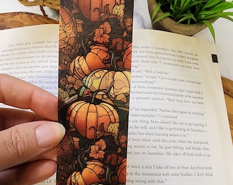 Puede incluir: Un marcapáginas decorativo con un diseño de calabaza. El marcapáginas tiene un fondo oscuro con calabazas naranjas y enredaderas verdes. Está hecho de un material resistente y tiene una borla naranja.