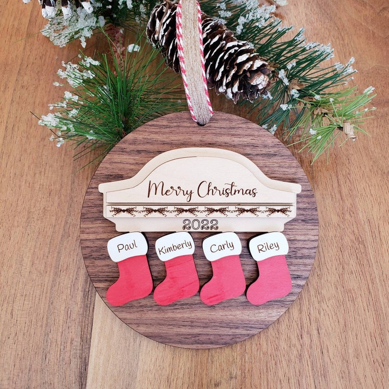 May include: A wooden Christmas ornament with a car silhouette and the text "Merry Christmas 2022".  Four stockings are attached to the bottom with the names "Paul", "Kimberly", "Carly", and "Riley" written on them.
