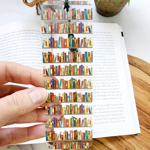 May include: A clear bookmark with a colorful design of a bookshelf filled with books. The bookmark is made of acrylic and has a rounded top.