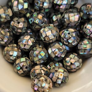 May include: A collection of round beads with a mosaic-like pattern. The beads are primarily dark gray with iridescent squares of abalone shell, reflecting colors of green, blue, and pink. The beads are in a white bowl.