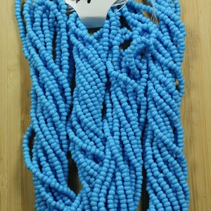 Peut inclure: Un ensemble de perles de rocaille bleu turquoise opaque. Les perles sont enfil&eacute;es ensemble, formant plusieurs brins. Une &eacute;tiquette attach&eacute;e aux perles indique "Blue Turquoise opaque". Les perles sont pr&eacute;sent&eacute;es sur une surface en bois brun clair.