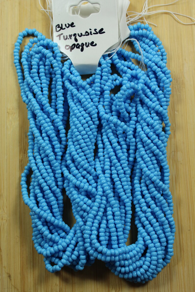Peut inclure: Un ensemble de perles de rocaille bleu turquoise opaque. Les perles sont enfil&eacute;es ensemble, formant plusieurs brins. Une &eacute;tiquette attach&eacute;e aux perles indique "Blue Turquoise opaque". Les perles sont pr&eacute;sent&eacute;es sur une surface en bois brun clair.