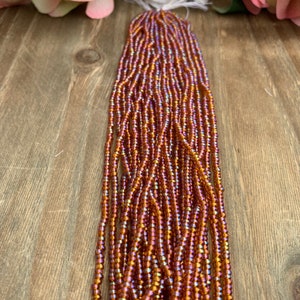 May include: Strands of iridescent orange and purple seed beads. The beads are strung together, creating a flowing, textured appearance. The beads are displayed on a wooden surface, with pink and white flowers in the background.