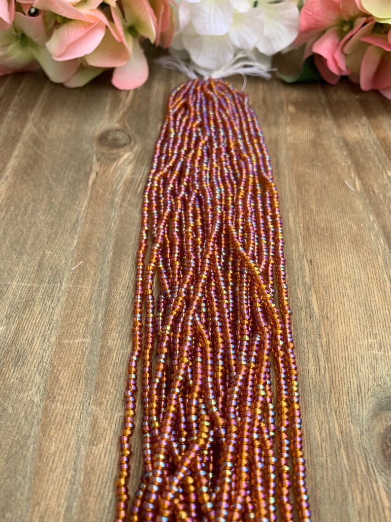 May include: Strands of iridescent orange and purple seed beads. The beads are strung together, creating a flowing, textured appearance. The beads are displayed on a wooden surface, with pink and white flowers in the background.