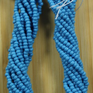 Peut inclure: Plusieurs brins de perles opaques turquoise, &eacute;troitement tiss&eacute;es. Les perles sont d'un bleu vif. Une &eacute;tiquette indique "Turquoise Opaque" au marqueur noir. Les perles sont pr&eacute;sent&eacute;es sur un fond en bois.
