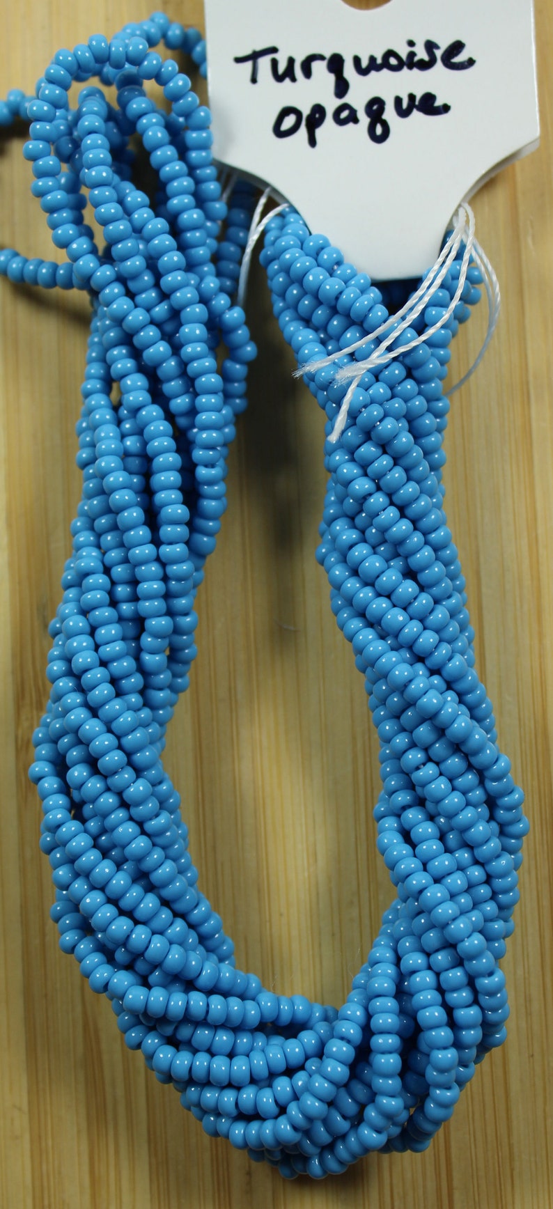 Peut inclure: Plusieurs brins de perles opaques turquoise, &eacute;troitement tiss&eacute;es. Les perles sont d'un bleu vif. Une &eacute;tiquette indique "Turquoise Opaque" au marqueur noir. Les perles sont pr&eacute;sent&eacute;es sur un fond en bois.