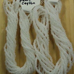 Peut inclure: Plusieurs brins de perles blanches, probablement pour la cr&eacute;ation de bijoux, sont pr&eacute;sent&eacute;s sur un fond en bois. Les perles sont petites et brillantes. Le texte "White pearl Ceylon" est visible sur une &eacute;tiquette.