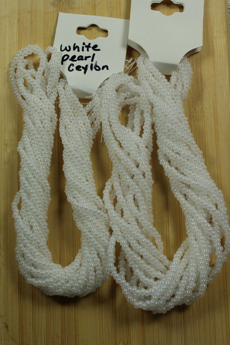 Peut inclure: Plusieurs brins de perles blanches, probablement pour la cr&eacute;ation de bijoux, sont pr&eacute;sent&eacute;s sur un fond en bois. Les perles sont petites et brillantes. Le texte "White pearl Ceylon" est visible sur une &eacute;tiquette.