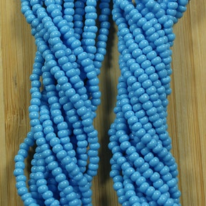 Peut inclure: Plusieurs brins de perles turquoise bleu clair opaques. Les petites perles rondes sont enfil&eacute;es ensemble. Une &eacute;tiquette indique "Blue Turquoise opaque". Les perles sont pr&eacute;sent&eacute;es sur une surface en bois.
