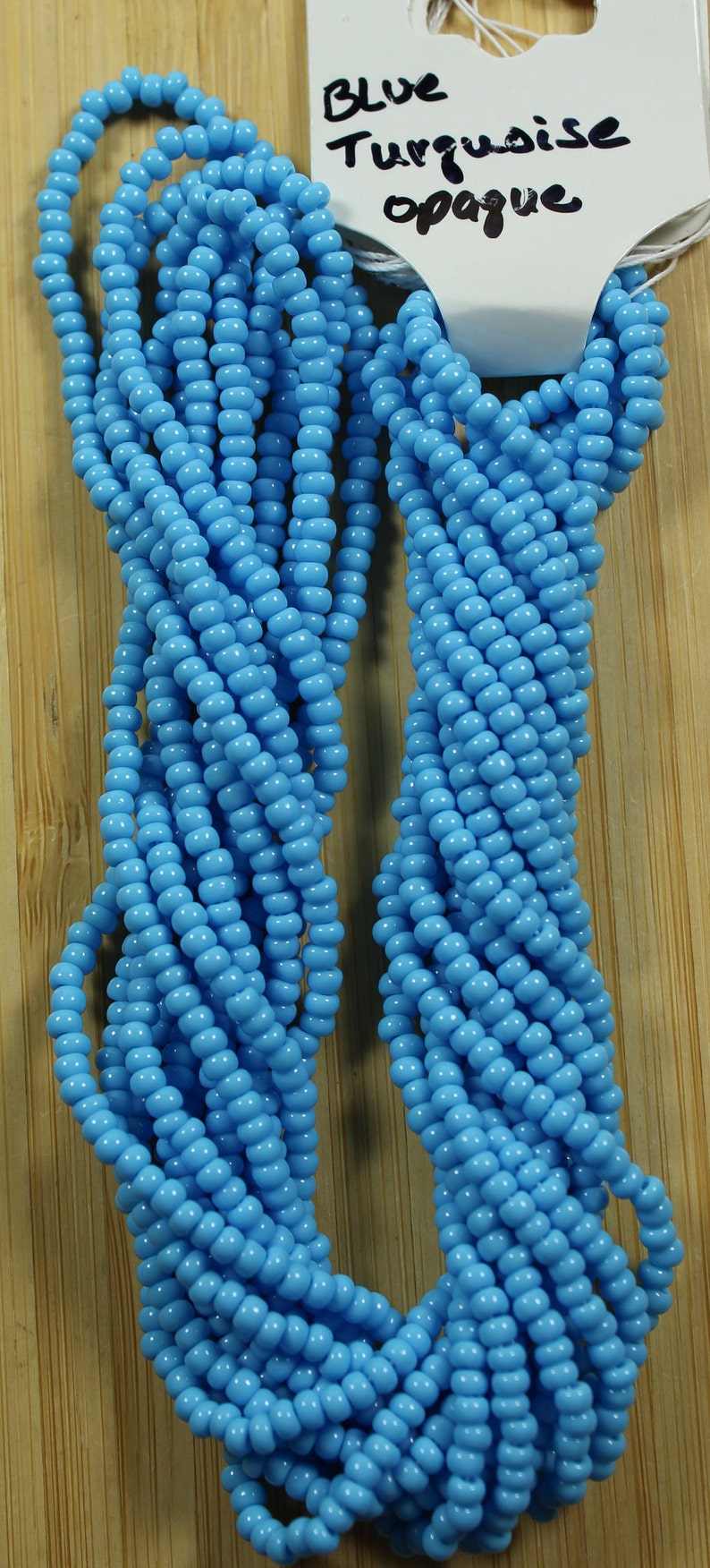 Peut inclure: Plusieurs brins de perles turquoise bleu clair opaques. Les petites perles rondes sont enfil&eacute;es ensemble. Une &eacute;tiquette indique "Blue Turquoise opaque". Les perles sont pr&eacute;sent&eacute;es sur une surface en bois.