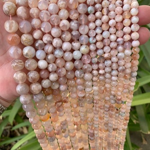 May include: Strands of faceted, round gemstone beads in varying sizes. The beads display a mix of peach, cream, and light brown hues, with subtle variations in color and pattern. The beads are strung together, ready for jewelry making.