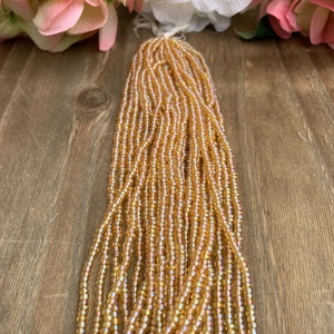 May include: Strands of small, iridescent, golden-colored seed beads. The beads are strung together, forming a thick bundle. The background includes pink and white flowers on a wooden surface.