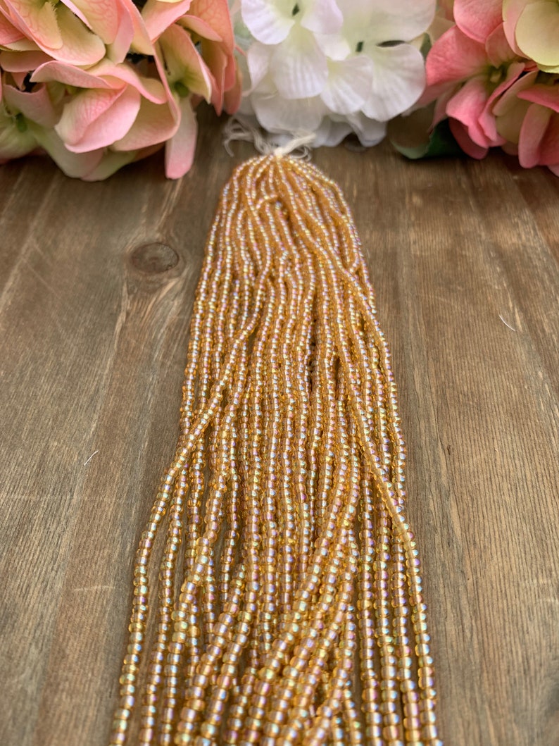 May include: Strands of small, iridescent, golden-colored seed beads. The beads are strung together, forming a thick bundle. The background includes pink and white flowers on a wooden surface.