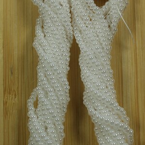 Peut inclure: Plusieurs rang&eacute;es de perles blanches, &eacute;tiquet&eacute;es "White pearl Ceylon", sont pr&eacute;sent&eacute;es sur un fond en bois brun clair. Les perles sont &eacute;troitement enfil&eacute;es, cr&eacute;ant un aspect textur&eacute; et fluide. L'image met en valeur la couleur et la texture des perles.