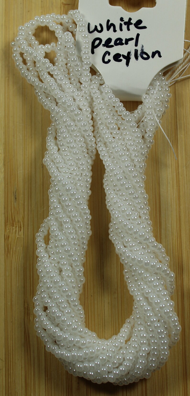 Peut inclure: Plusieurs rang&eacute;es de perles blanches, &eacute;tiquet&eacute;es "White pearl Ceylon", sont pr&eacute;sent&eacute;es sur un fond en bois brun clair. Les perles sont &eacute;troitement enfil&eacute;es, cr&eacute;ant un aspect textur&eacute; et fluide. L'image met en valeur la couleur et la texture des perles.