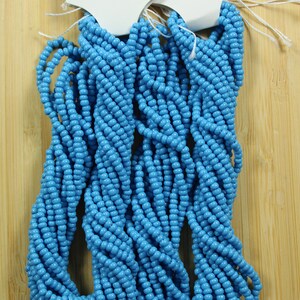 Peut inclure: Plusieurs brins de perles de rocaille opaques turquoise. Les perles sont d'un bleu vif et sont enfil&eacute;es ensemble. Les perles sont pr&eacute;sent&eacute;es sur une surface en bois brun clair. Les &eacute;tiquettes fix&eacute;es aux perles indiquent "Turquoise Opaque".