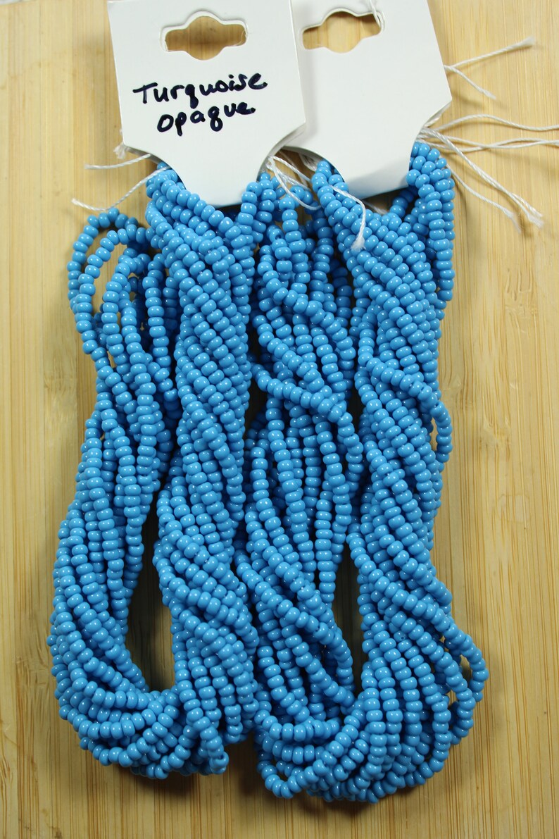 Peut inclure: Plusieurs brins de perles de rocaille opaques turquoise. Les perles sont d'un bleu vif et sont enfil&eacute;es ensemble. Les perles sont pr&eacute;sent&eacute;es sur une surface en bois brun clair. Les &eacute;tiquettes fix&eacute;es aux perles indiquent "Turquoise Opaque".