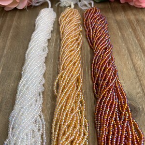 May include: Three strands of seed beads in white, gold, and reddish-brown hues. The beads are strung together, creating a textured, rope-like appearance. The beads are iridescent, reflecting light.