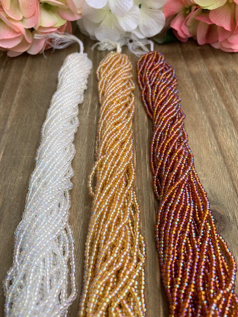 May include: Three strands of seed beads in white, gold, and reddish-brown hues. The beads are strung together, creating a textured, rope-like appearance. The beads are iridescent, reflecting light.