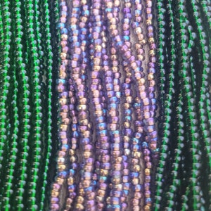 May include: Close-up of strands of beads in shades of green and iridescent purple. The beads are strung together, creating a textured and colorful display. The beads are small and round, and the overall effect is eye-catching.