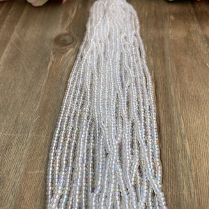 May include: A bundle of iridescent, clear glass seed beads. The beads are strung together, creating multiple strands. The beads have a subtle, shimmering effect, reflecting light. The background features a wooden surface and soft pink and white flowers.