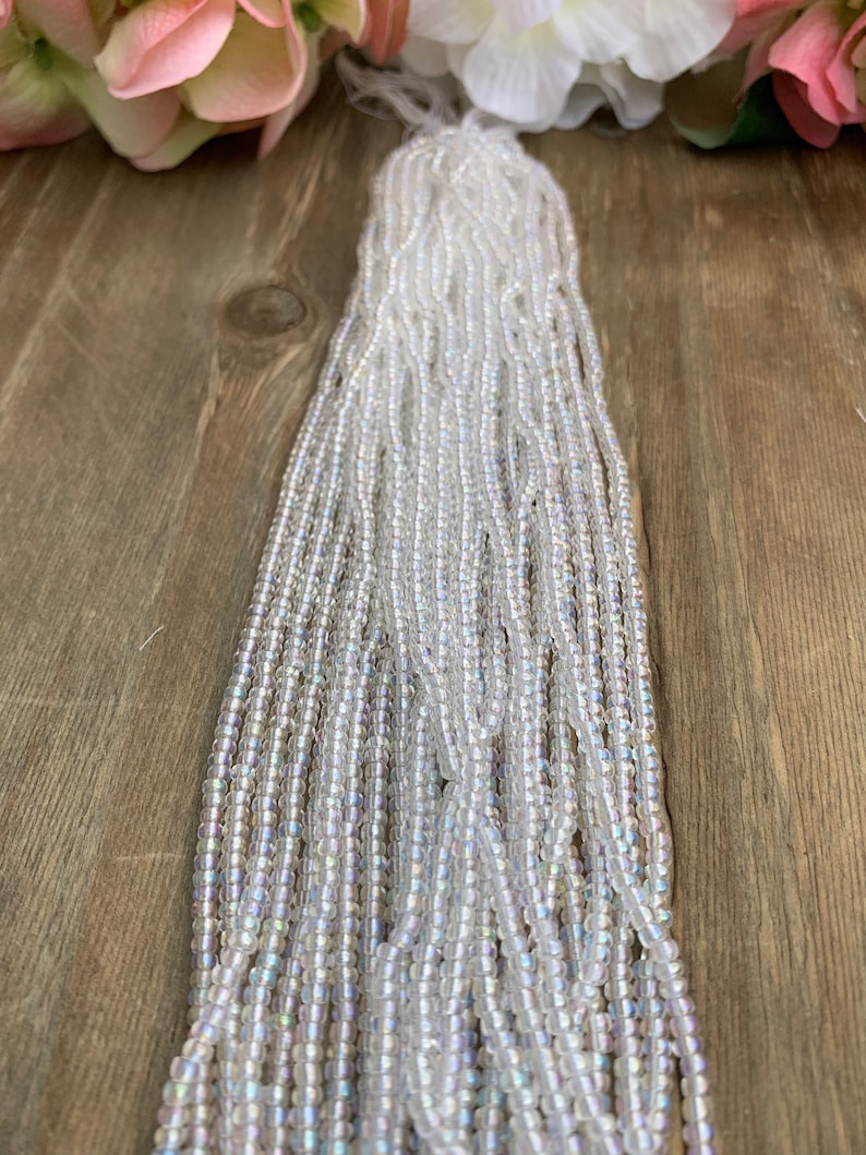 May include: A bundle of iridescent, clear glass seed beads. The beads are strung together, creating multiple strands. The beads have a subtle, shimmering effect, reflecting light. The background features a wooden surface and soft pink and white flowers.