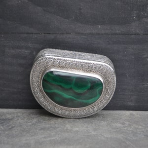 May include: A silver trinket box with an oval-shaped green malachite stone set in the center. The box is decorated with a repeating pattern of small circles and dots.
