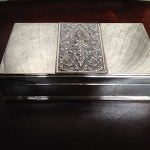 May include: A rectangular box with a silver-colored metal frame and a light-colored, wood-grain top. The center of the lid features an ornate silver panel with a detailed figure and floral designs. The box is closed and sits on a dark surface.