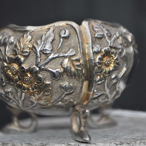 May include: A silver bowl with intricate floral designs and three legs. The bowl is decorated with raised flowers and leaves, and the metal has a slightly gold tone.