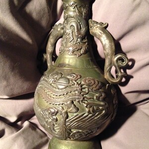 May include: An antique bronze vase with intricate dragon and phoenix relief carvings. The vase features two dragon-shaped handles and detailed patterns. The color is a mix of bronze and silver, with a weathered appearance, showcasing its age and craftsmanship.