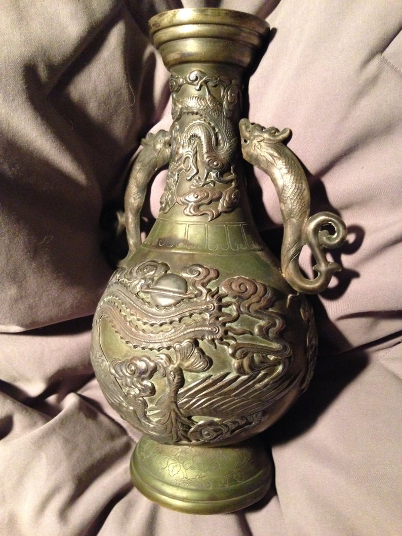 May include: An antique bronze vase with intricate dragon and phoenix relief carvings. The vase features two dragon-shaped handles and detailed patterns. The color is a mix of bronze and silver, with a weathered appearance, showcasing its age and craftsmanship.