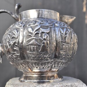 May include: A silver pitcher with an ornate design featuring a house and a figure. The pitcher has a handle and a spout.