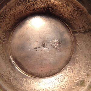 May include: Close-up of a silver-toned bowl interior. The bowl's interior has a circular design with a central indentation. The rim features a floral pattern. There are two small markings near the center of the bowl.