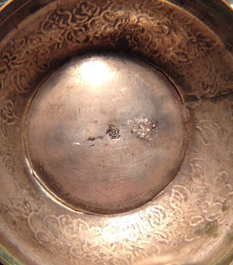 May include: Close-up of a silver-toned bowl interior. The bowl's interior has a circular design with a central indentation. The rim features a floral pattern. There are two small markings near the center of the bowl.