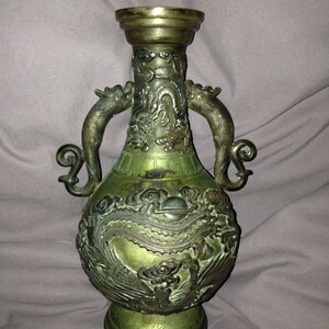 May include: An antique bronze vase with intricate dragon and cloud relief carvings. The vase features two dragon-shaped handles and a flared rim. The overall color is a mix of bronze and green, with a detailed base.