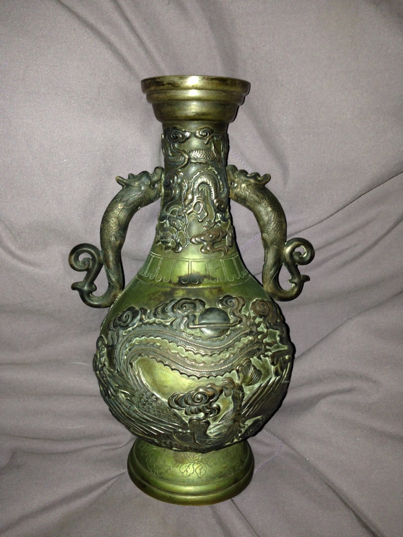 May include: An antique bronze vase with intricate dragon and cloud relief carvings. The vase features two dragon-shaped handles and a flared rim. The overall color is a mix of bronze and green, with a detailed base.