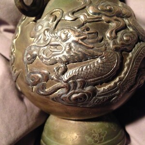 May include: Antique bronze-colored vase with intricate dragon and cloud relief carvings. The vase has a bulbous body, a flared base, and a curved handle. The dragon design is highly detailed, with scales and flowing features.