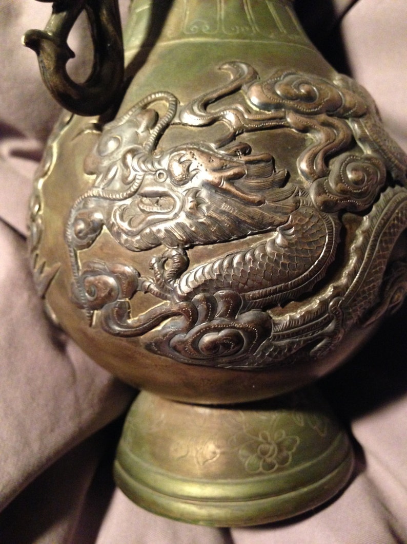 May include: Antique bronze-colored vase with intricate dragon and cloud relief carvings. The vase has a bulbous body, a flared base, and a curved handle. The dragon design is highly detailed, with scales and flowing features.