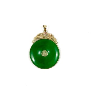 May include: A round, vibrant green jade pendant with a gold-colored filigree design. The pendant features a small, red gemstone and a circular emblem in the center. The jade is approximately 2 inches in diameter. The pendant is suspended from a gold-colored bail.