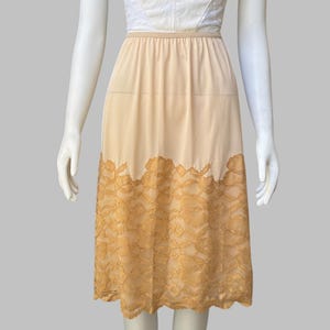 May include: A white lace-trimmed bodice top paired with a beige knee-length skirt featuring an elastic waistband and a wide band of tan floral lace along the bottom hem. The garment is displayed on a mannequin against a neutral gray background.
