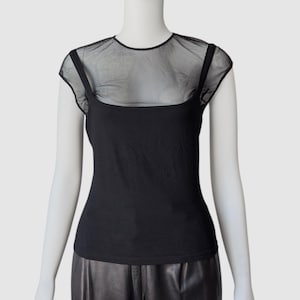 May include: Black top with a sheer mesh overlay. The top features a square neckline and thin straps. The mesh covers the shoulders and upper chest. The garment is displayed on a mannequin.
