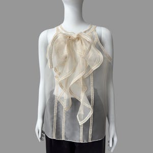 May include: A sleeveless, sheer cream-colored top with a large, ruffled bow detail at the neckline. The top features vertical, embroidered lines and is displayed on a mannequin. The garment is a delicate, elegant piece.