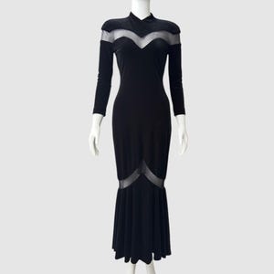 以下が含まれることがあります： ハイネックとマーメイドヘムが特徴の黒の長袖ドレス。胸元とスカートの裾にシアーメッシュパネルが施されています。白いマネキンに着せられたドレスです。