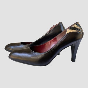 Pode incluir: Sapatos de salto alto de couro preto com detalhe de cobra na parte de trás. Os sapatos têm bico fino e salto alto.