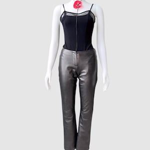 May include: A black bodysuit with spaghetti straps and a sheer neckline is paired with metallic gray leather pants. A red floral necklace adds a pop of color to the outfit. The ensemble is displayed on a mannequin.