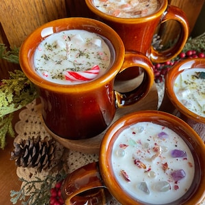 May include: Several brown ceramic mug candles with white wax, decorated with crystals, herbs, and a candy cane. The mugs have a glossy finish and are arranged on a wooden surface, creating a cozy, festive atmosphere.