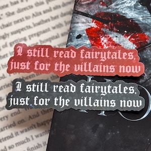 May include: Two red and black bookmarks with white text. The top bookmark reads 'I still read fairytales, just for the villains now.' The bottom bookmark reads 'I still read fairytales, just for the villains now.' The bookmarks are placed on a black book cover with a clock face design. The clock face has a white background with black hands and black roman numerals. The clock face is surrounded by a dark gray background with a red and black galaxy design.