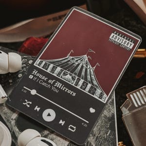 May include: A black and white music player sticker with a red background. The sticker has a graphic of a circus tent and the text 'House of Mirrors' and 'If I Catch You'. The sticker has a play button, a heart icon, and a timer that reads 0:30 and 4:20. The sticker is on a black surface with a gold lighter, a book, and red rose petals.