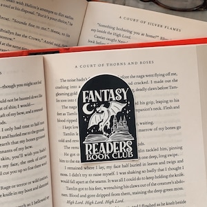 以下が含まれることがあります： 黒と白のファンタジー読書クラブステッカー。ドラゴンと城が描かれています。ステッカーは、「A Court of Thorns and Roses」というタイトルの表紙のついた本に貼られています。