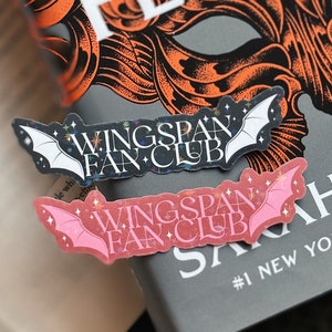 以下が含まれることがあります： コウモリのデザインと「Wingspan Fan Club」という文字が白で書かれた黒とピンクの背景のステッカー2枚。ステッカーは、「Six of Crows」と「#1 New York」という文字が書かれた本の表紙に貼られています。